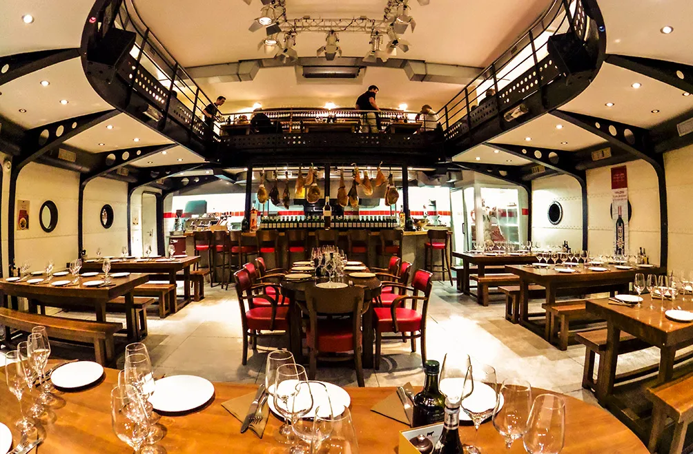 Interior of a traditional Bordeaux brasserie featuring wooden tables with red chairs, a central bar with wine bottles, exposed black beam ceiling, and warm lighting throughout the dining room.