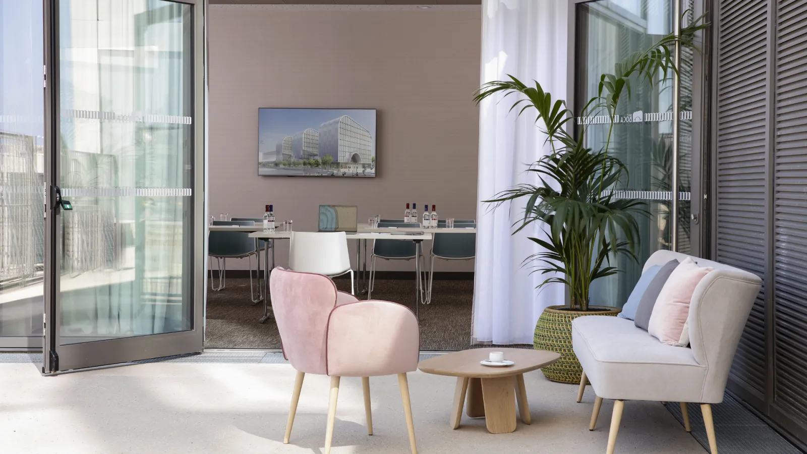 Modern hotel suite at Hilton Garden Inn Bordeaux Centre featuring a bright lounge area with contemporary furniture, a pink accent chair, beige sofa, wooden side table, large windows overlooking the city, and a work desk in the background