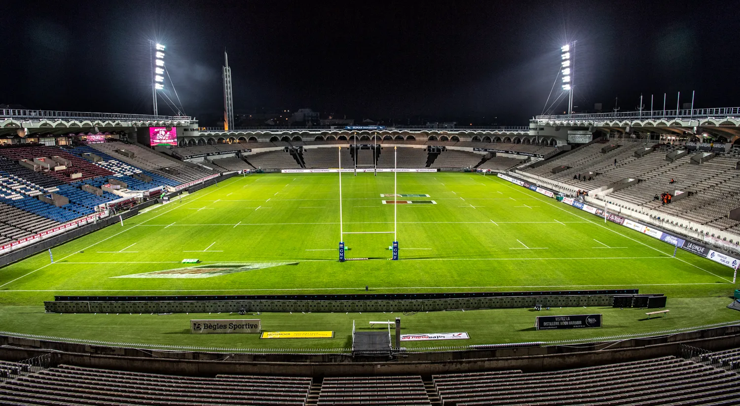 Stade Matmut Bordeaux rugby field at night, illuminated with floodlights, showing the white lines of the rugby pitch with two goal posts centered, surrounded by tiered seating in the stadium.