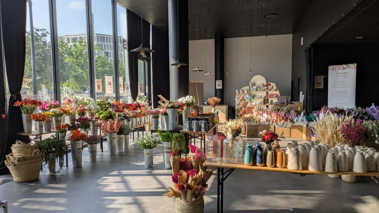 A bright flower shop interior in Bordeaux displaying numerous fresh and dried flowers in vases, ceramic vessels, and arrangements on tables and shelves, with large windows overlooking the city and modern pendant lights overhead.