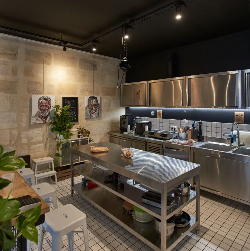 Professional kitchen interior with stainless steel equipment, central work table, white tile flooring, portrait artwork on beige walls, potted plants, and modern overhead lighting