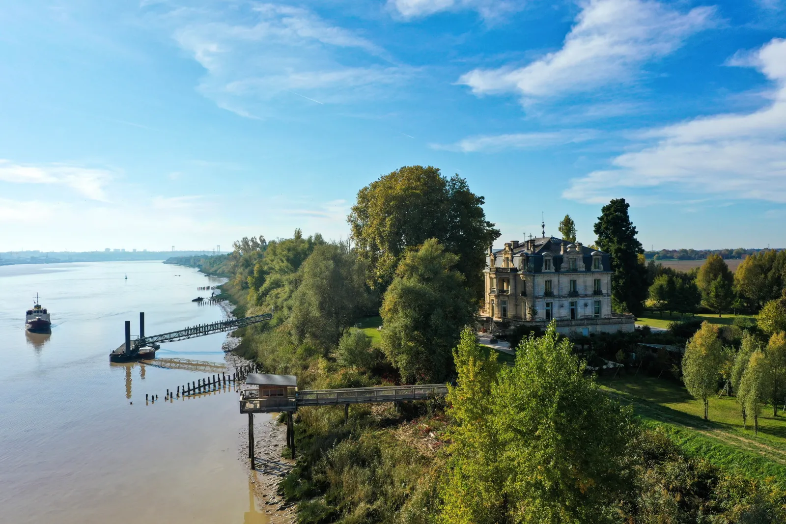 Aerial view of Château Grattequina overlooking the Garonne River in Bordeaux, surrounded by lush green vegetation and connected by a wooden bridge to the riverbank, with a cargo ship visible on the water.