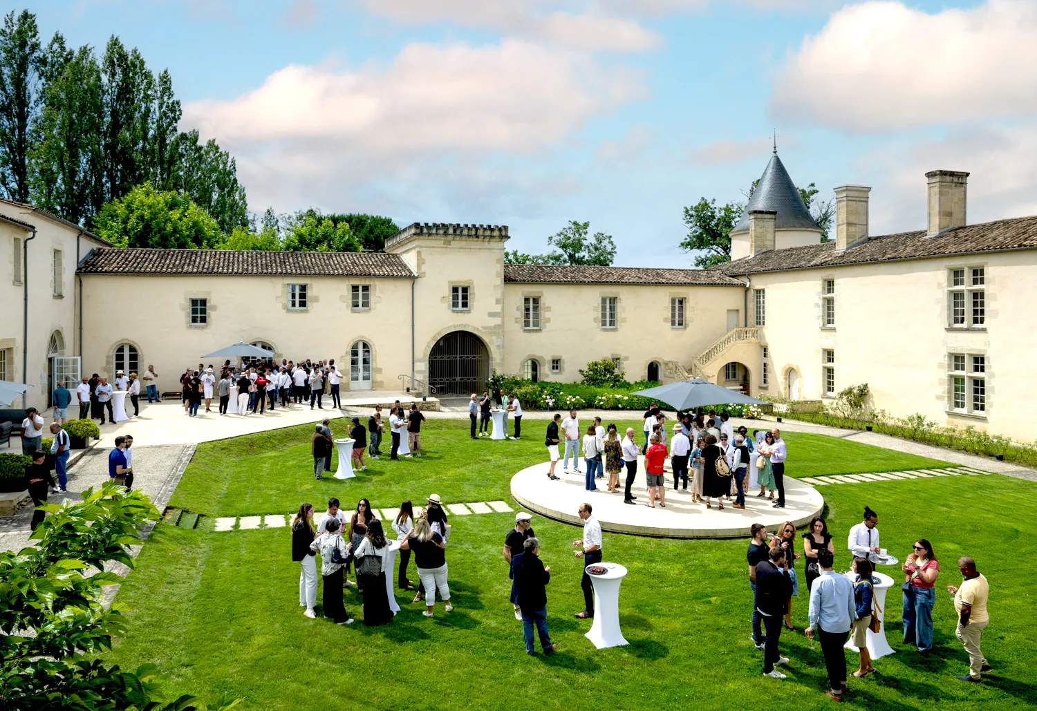 Guests mingling in the courtyard of Château Toulouse-Lautrec in Bordeaux during an outdoor event, with cream-colored château buildings, manicured lawns, and white decorative elements visible.