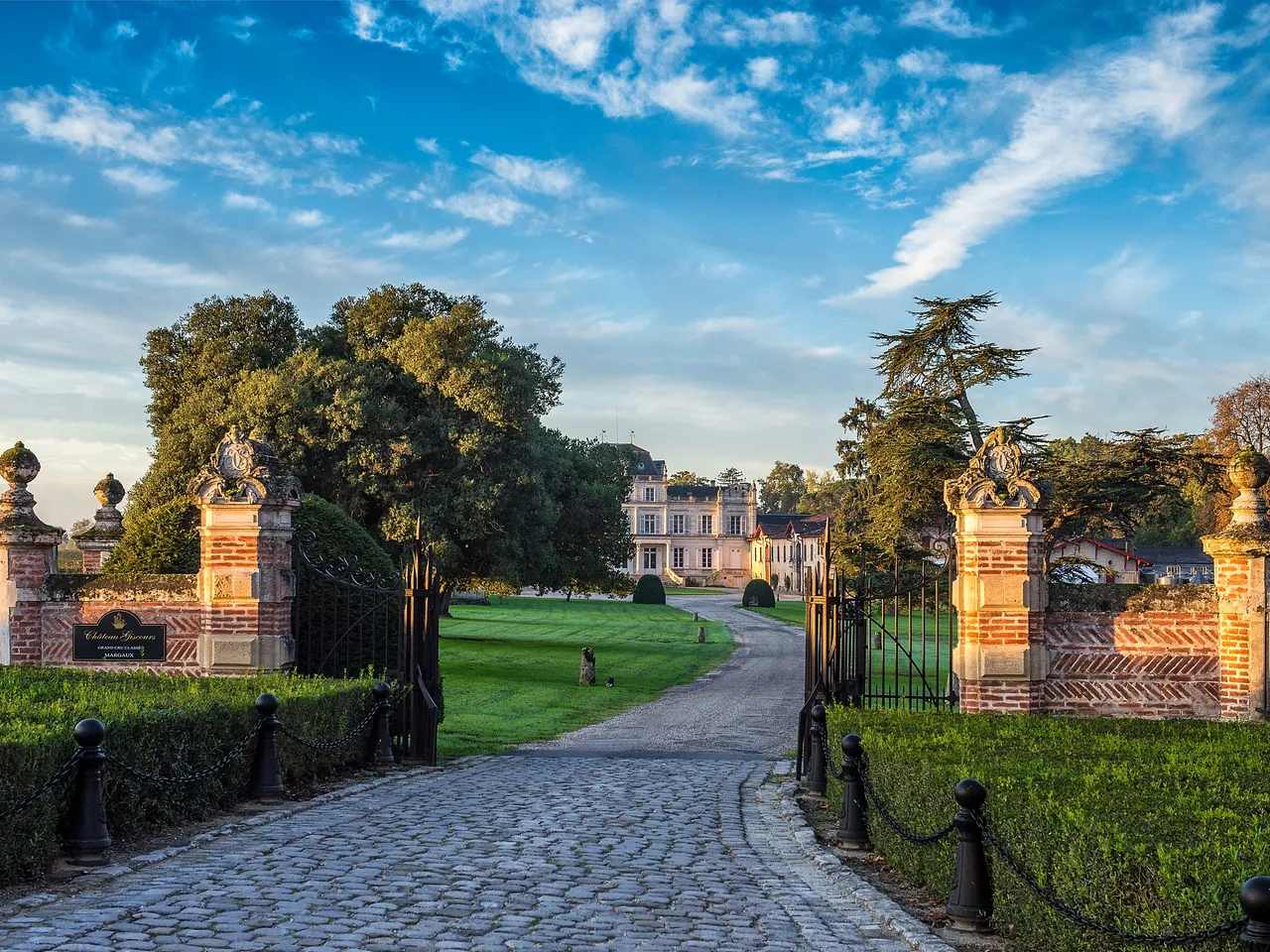 Grand entrance gates of Château Giscours in Bordeaux, with ornate stone pillars flanking a tree-lined driveway leading to a pink neoclassical mansion under a blue sky.