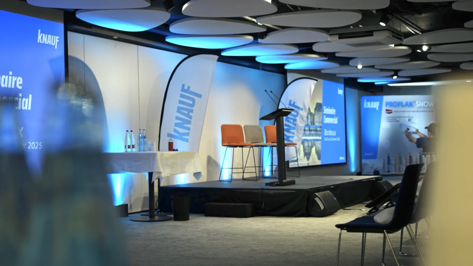 Modern conference hall with Knauf branding, featuring a stage with orange chairs, blue lighting panels on the ceiling, presentation screens, and an audience seating area