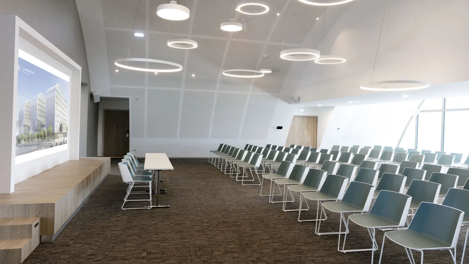 Modern conference room at Hilton Garden Inn Bordeaux Centre with rows of green chairs, a presentation screen displaying an architectural project, circular ceiling lights, and large windows overlooking the city.