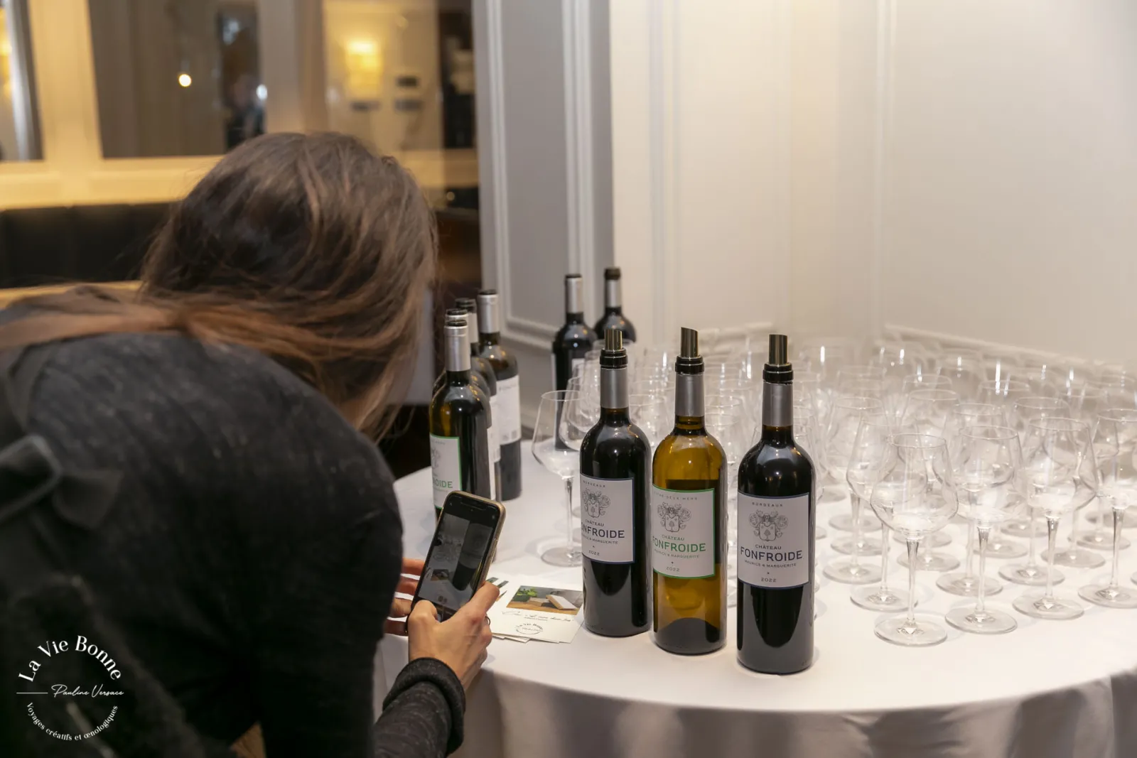 Woman photographing wine bottles from the Bonfroide vineyard displayed on a white table with empty glasses during a wine tasting event in Bordeaux