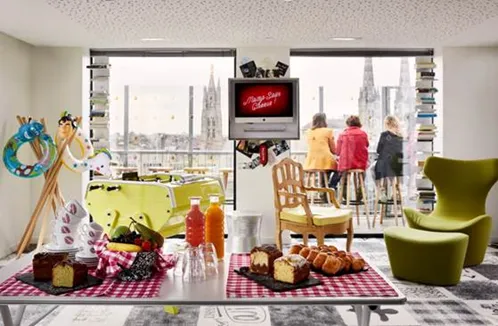 Colorful modern interior space at Mama Shelter Bordeaux with pink checkered table, fresh pastries and beverages, green armchairs, large windows overlooking the city skyline, and guests enjoying the vibrant atmosphere.