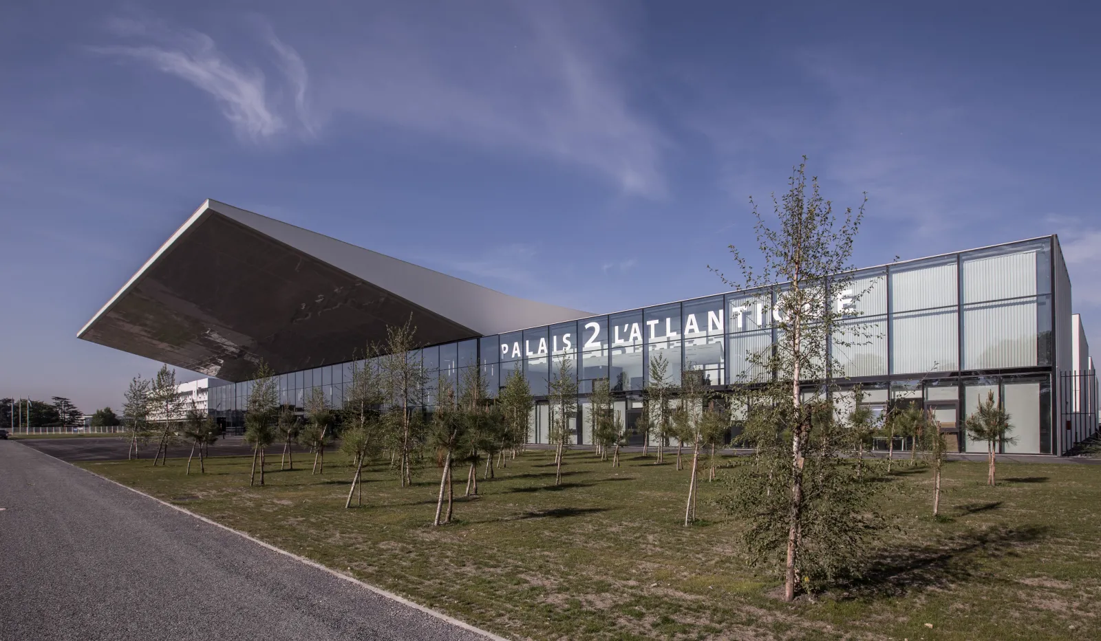 Modern glass and steel exhibition hall with 'Palais 2 L'Atlantique' signage, surrounded by young trees and landscaping in Bordeaux.