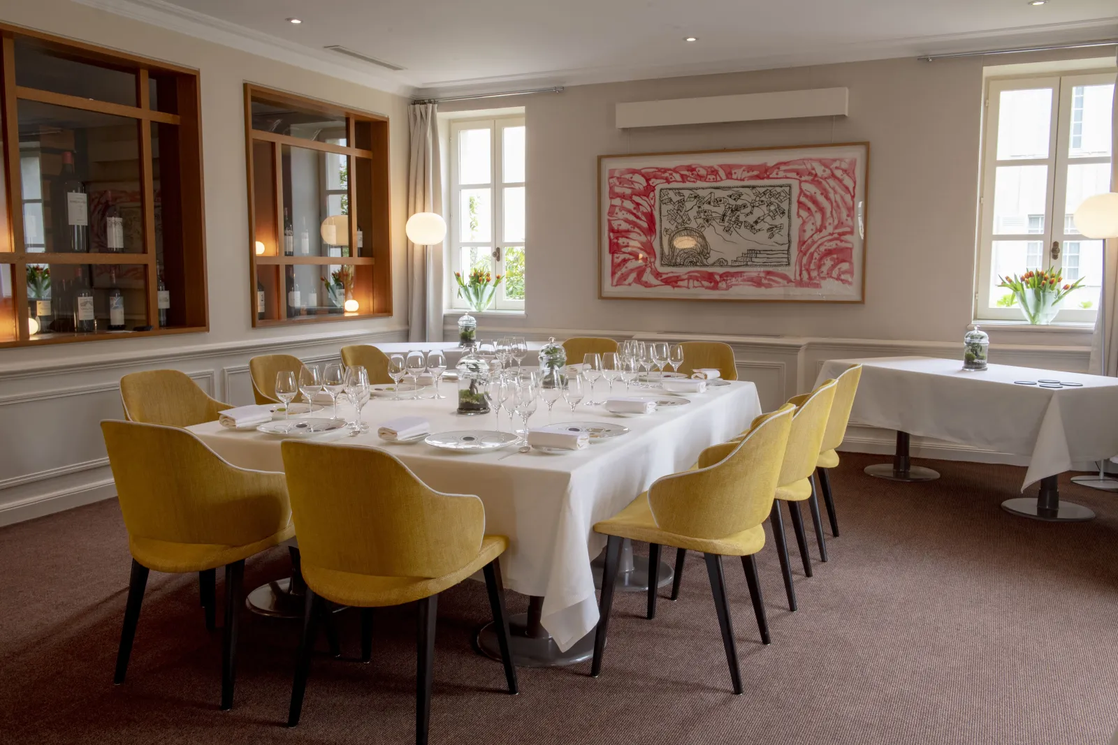 Elegant dining room with a rectangular table set for formal dinner, yellow upholstered chairs, white tablecloth, and place settings. The room features wooden display cabinets, framed artwork with red border, large windows with natural light, and beige wal