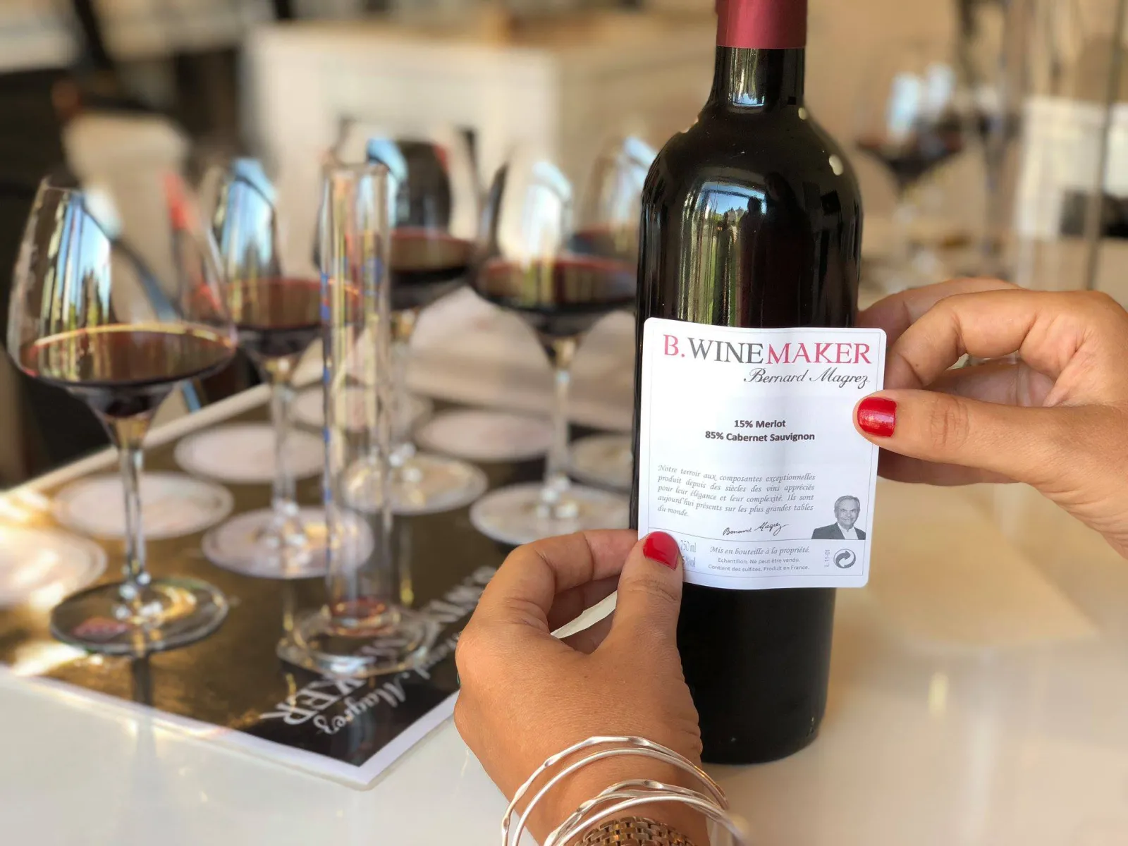 A person holding a bottle of B.Winemaker wine by Bernard Magrez, showing a blend of 15% Merlot and 85% Cabernet Sauvignon, with wine glasses and place settings visible on a dining table in the background.