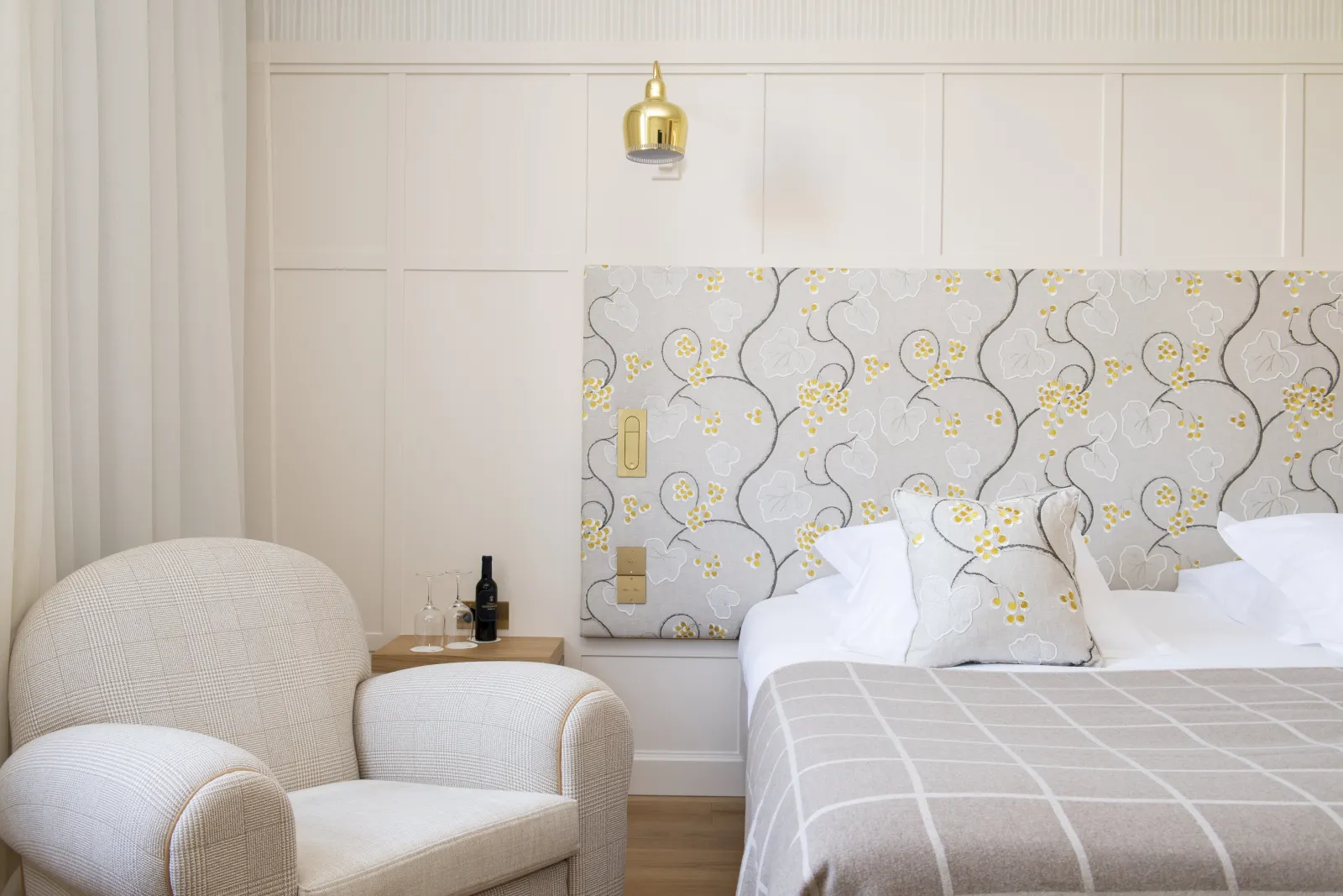 Modern bedroom at Como Cordeillan-Bages in Bordeaux featuring a bed with white linens, decorative headboard with yellow floral pattern, cream upholstered armchair, wooden nightstand, brass pendant light, and minimalist wall paneling