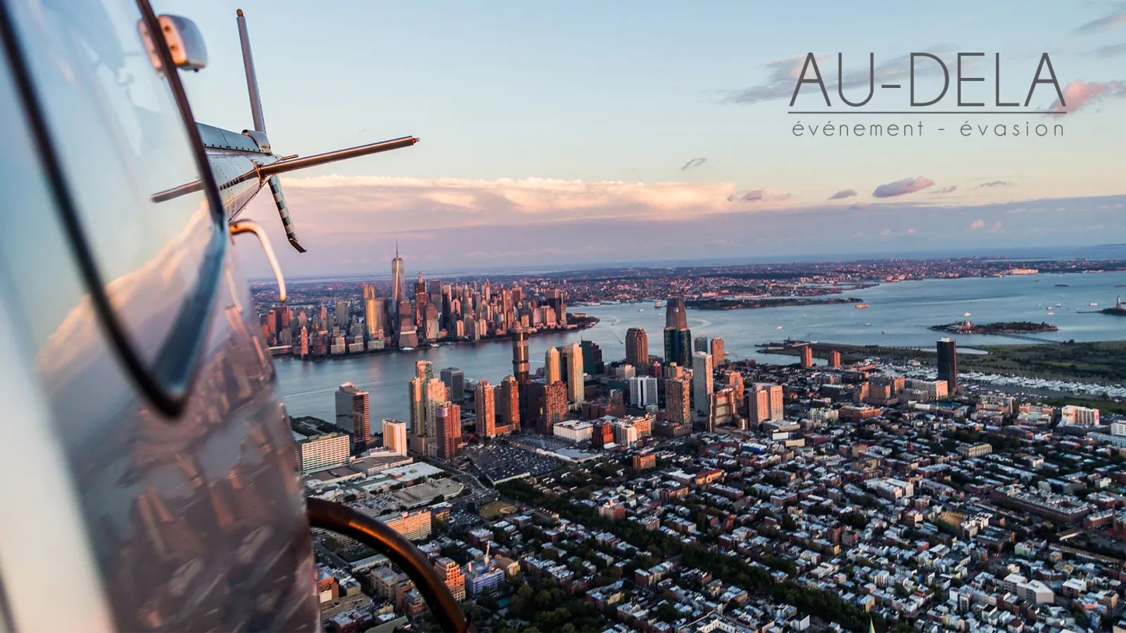 Aerial view of Manhattan skyline at sunset taken from a helicopter, showing skyscrapers, waterways, and surrounding boroughs with the aircraft propeller visible in the foreground.