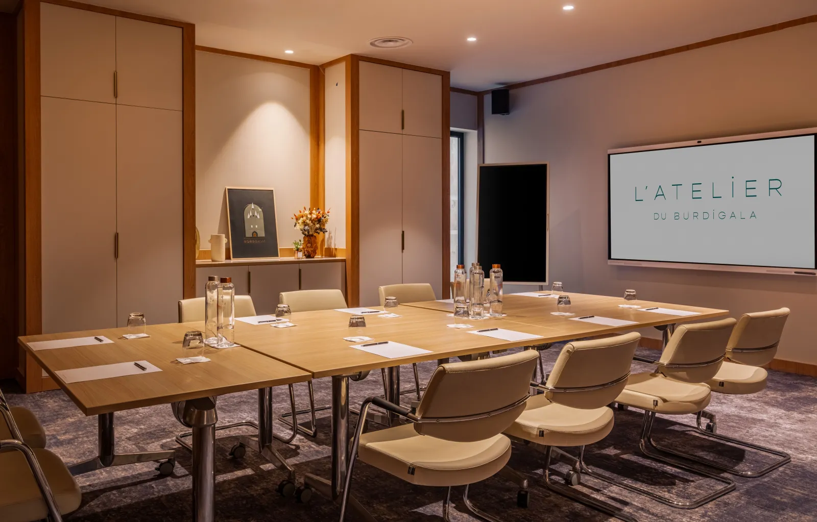 Modern conference room at Hôtel Burdigala with wooden U-shaped table, beige chairs, water glasses, projection screen displaying 'L'ATELIER au BURDIGALA', and elegant cabinetry with decorative accessories