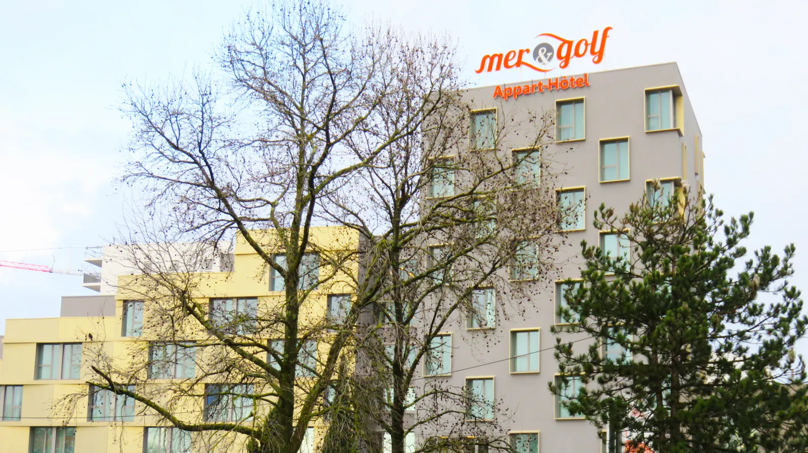 Exterior view of Mer & Golf Aparthotel, a modern multi-story building with beige and yellow facades, featuring regular window arrangements and the hotel signage on top, surrounded by bare trees in winter.