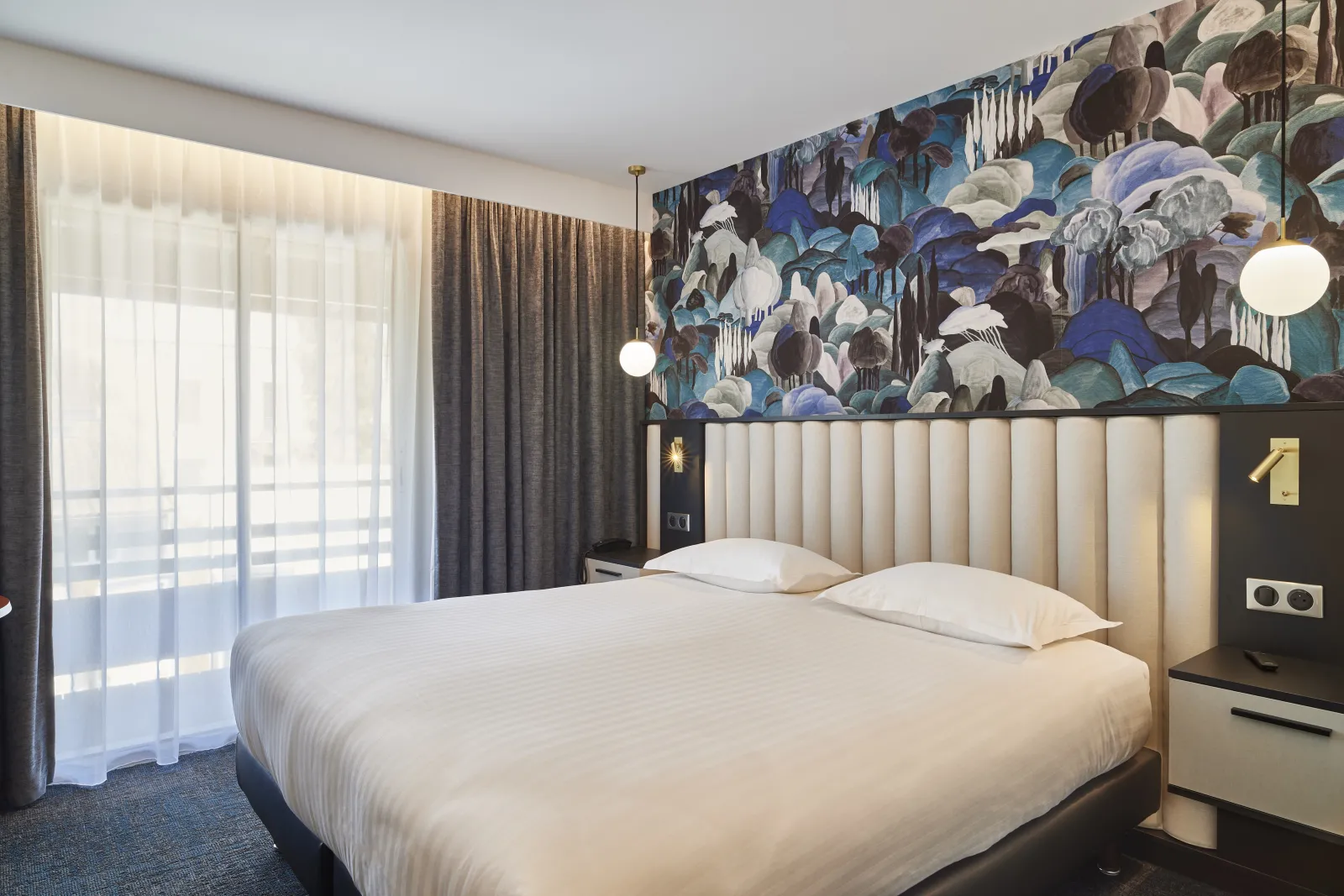 Modern hotel bedroom with a queen bed, cream upholstered headboard, dark accent wall with blue floral artwork, pendant lights, floor-to-ceiling windows with curtains, and contemporary furnishings at Best Western Premier Hôtel Des Vignes Et Des Anges in Bo
