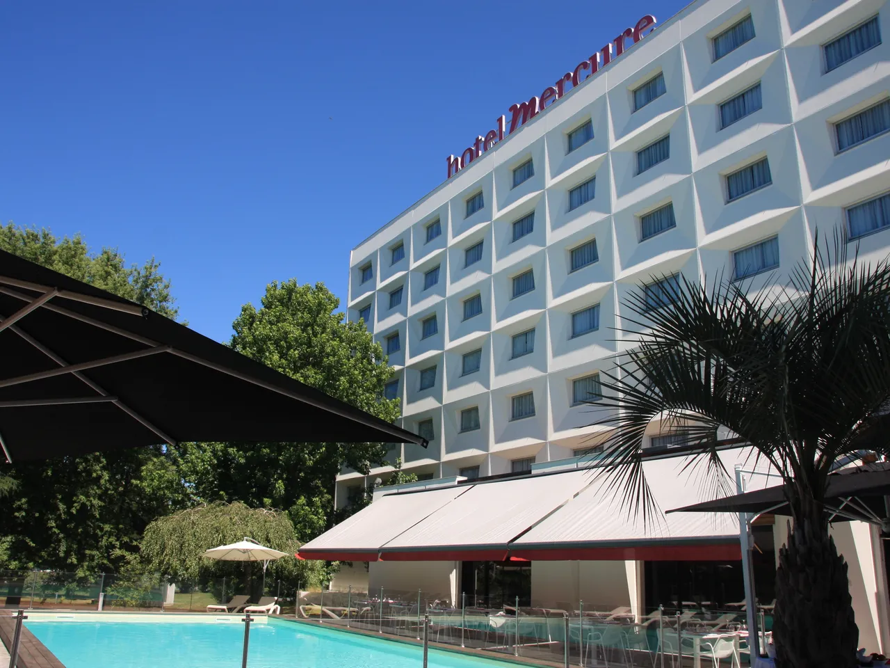 Mercure hotel building with white facade overlooking a swimming pool with red and white umbrellas and palm trees under blue sky in Bordeaux-Lac
