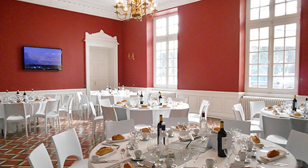 Elegant formal dining room with red walls, white chairs, round tables set with wine bottles and place settings, crystal chandelier, large windows, and flat-screen TV.
