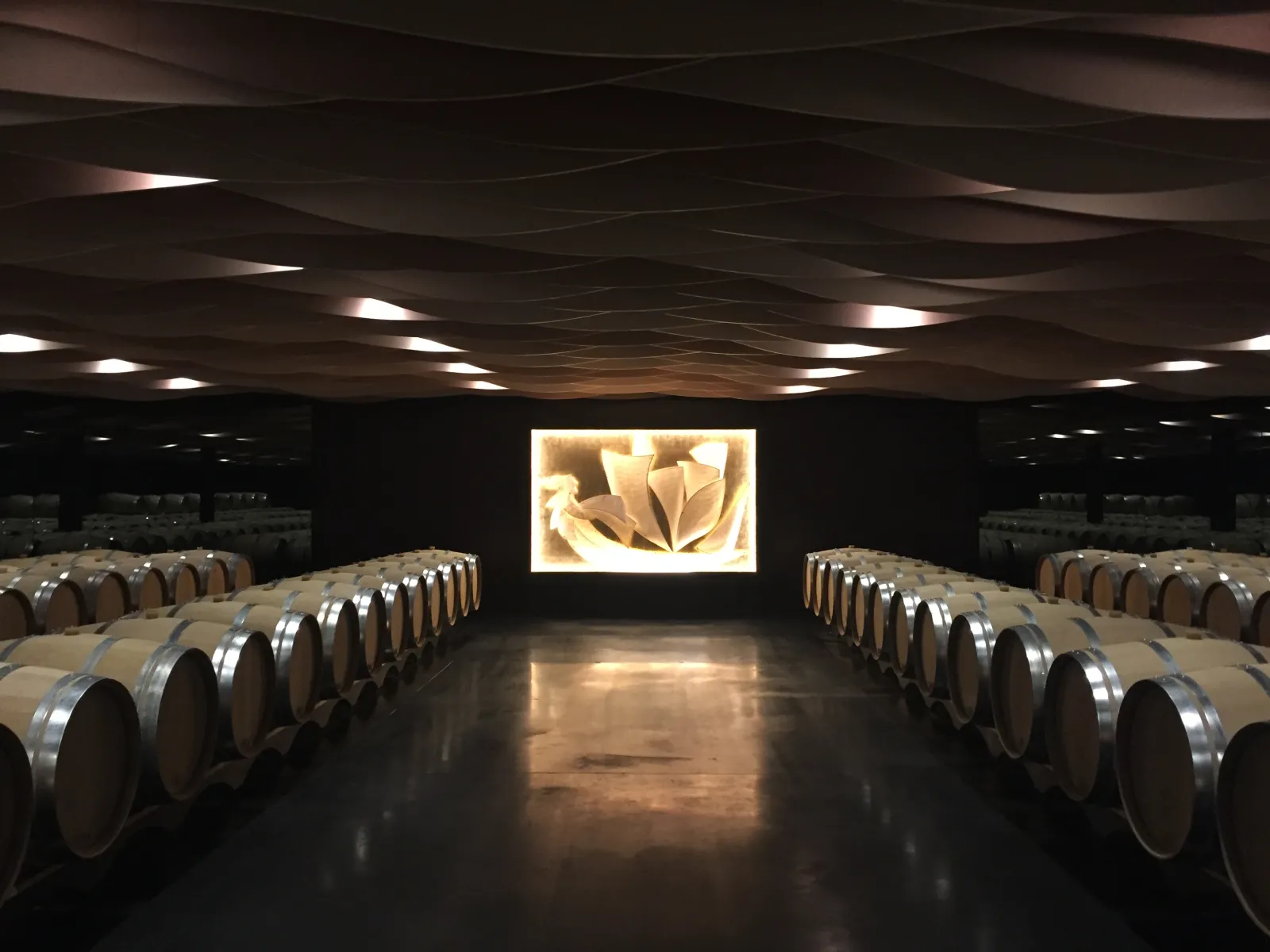 Modern wine cellar with wooden barrels lined symmetrically on both sides, polished concrete floor, wavy ceiling with integrated lighting, and a illuminated screen displaying an abstract wine-related image in Bordeaux.