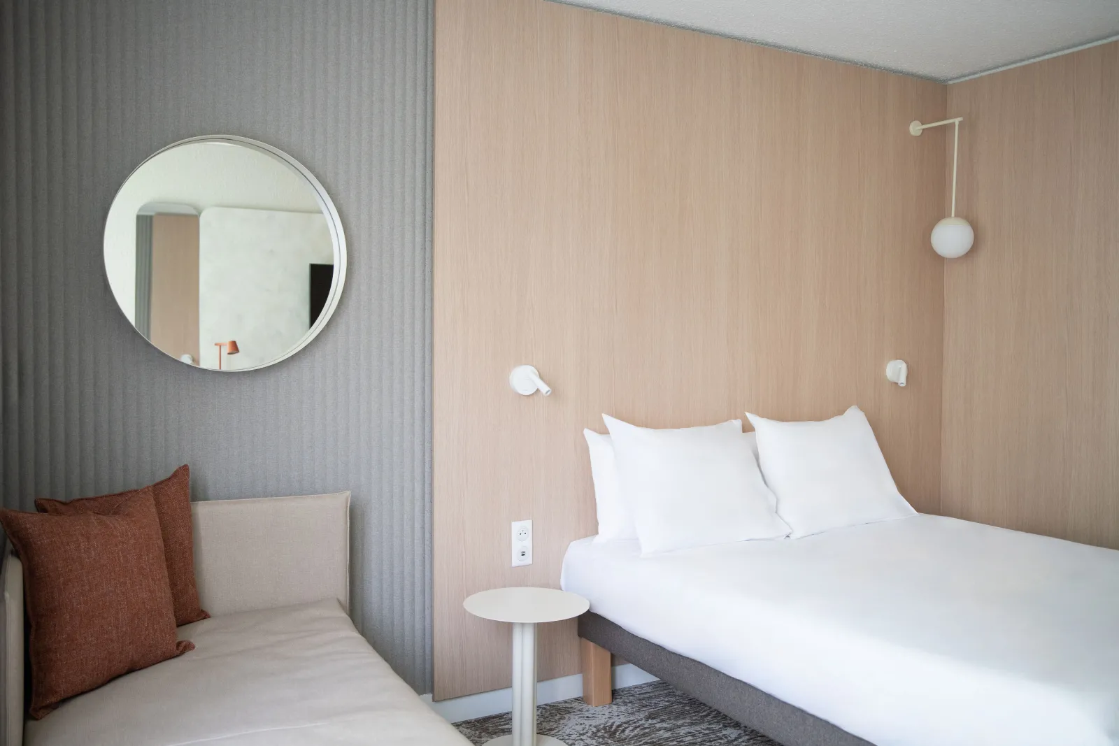 Modern hotel bedroom with a double bed, white bedding, round mirror, minimalist lighting fixtures, and warm wood paneling at Novotel Bordeaux Mérignac