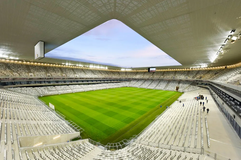 Interior view of Stade Atlantique Bordeaux Métropole, a modern football stadium with white seating, a bright green pitch, and a distinctive angular roof opening to the sky.