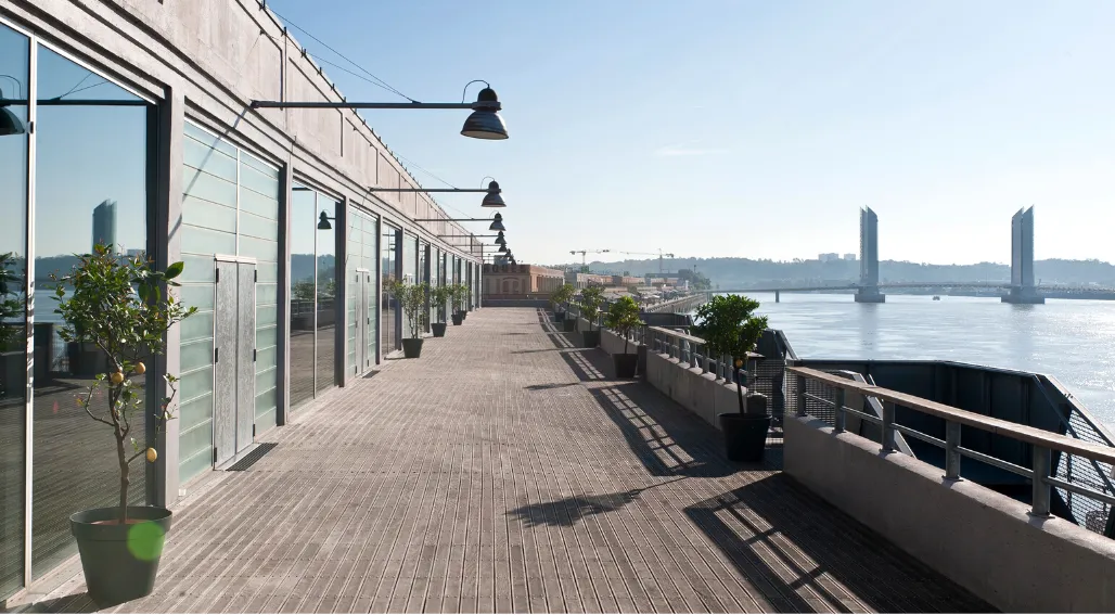 Modern waterfront terrace at Hangar 14 in Bordeaux, featuring a wooden deck with white buildings, black pendant lights, potted plants, and a view of the Garonne River with a bridge in the background under clear skies.