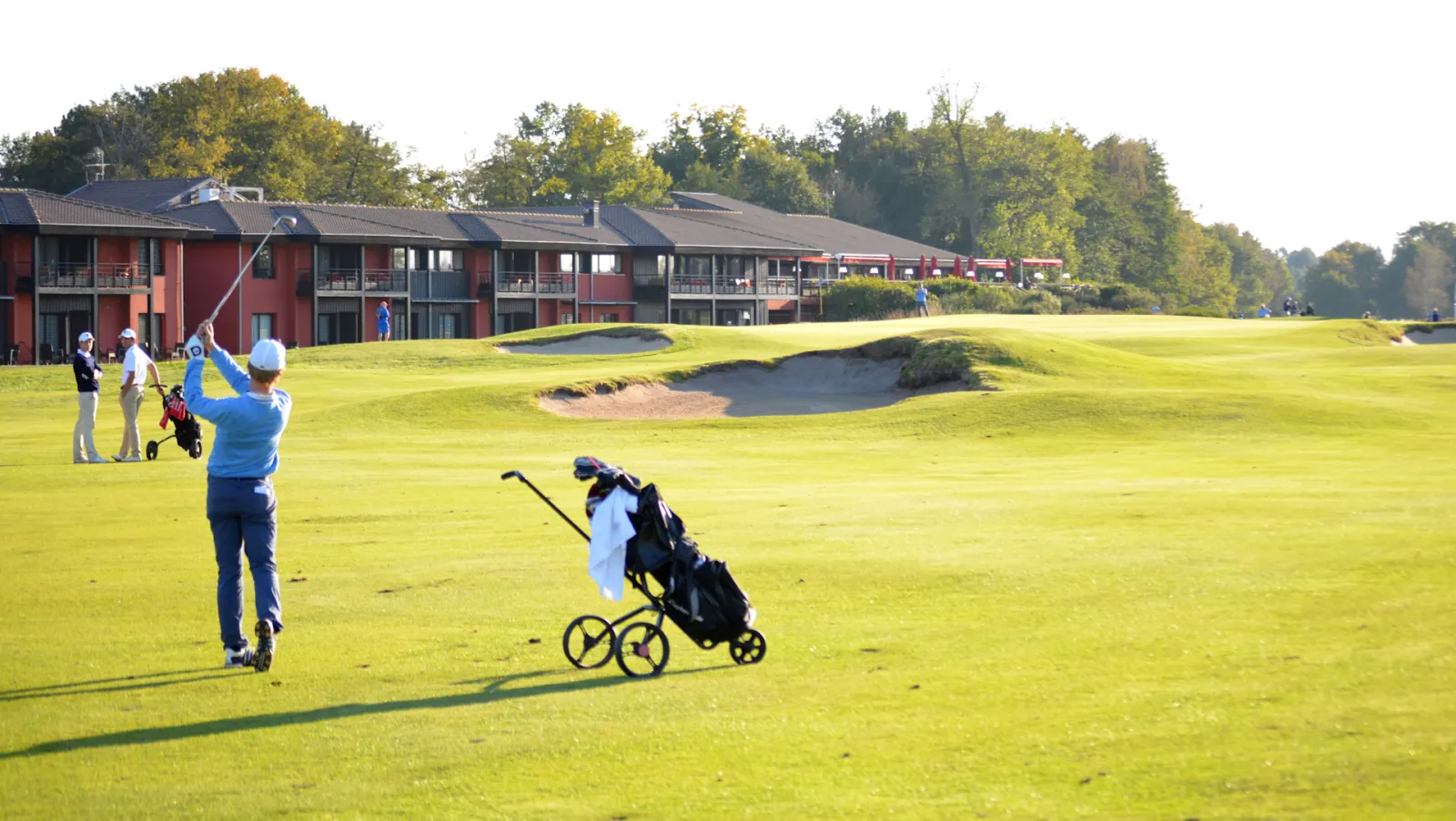 Golfers playing on a pristine championship golf course with modern red and black clubhouse buildings in the background, surrounded by manicured green fairways and sand bunkers.