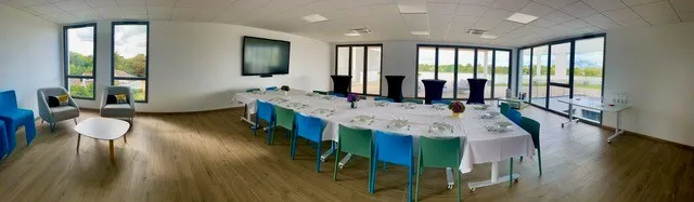Modern event space with blue chairs around a long white table, large windows overlooking the landscape, wooden flooring, and contemporary furnishings in a bright, airy room.