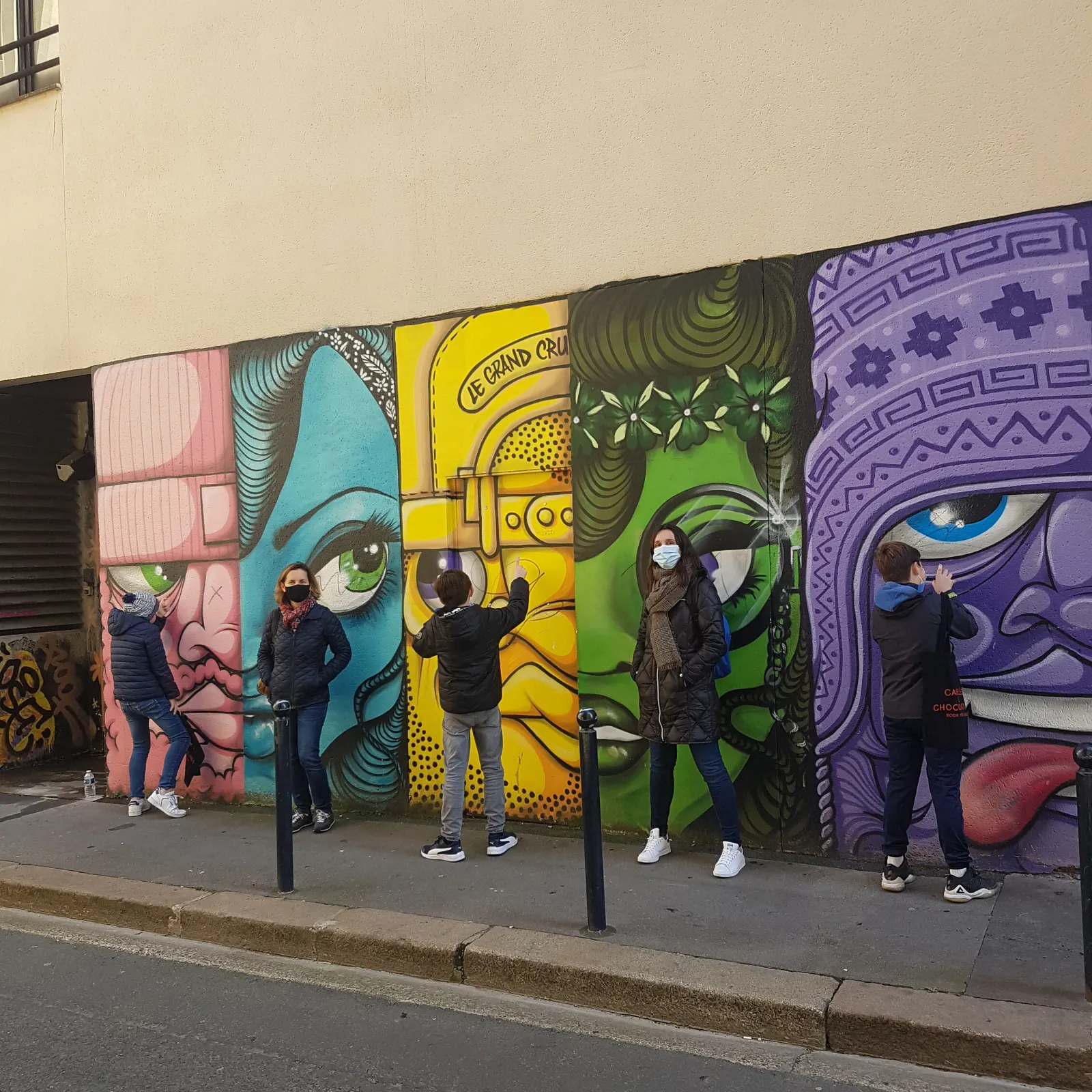 Five people posing in front of a colorful street art mural featuring stylized faces in pink, turquoise, yellow, green, and purple tones on a Bordeaux street