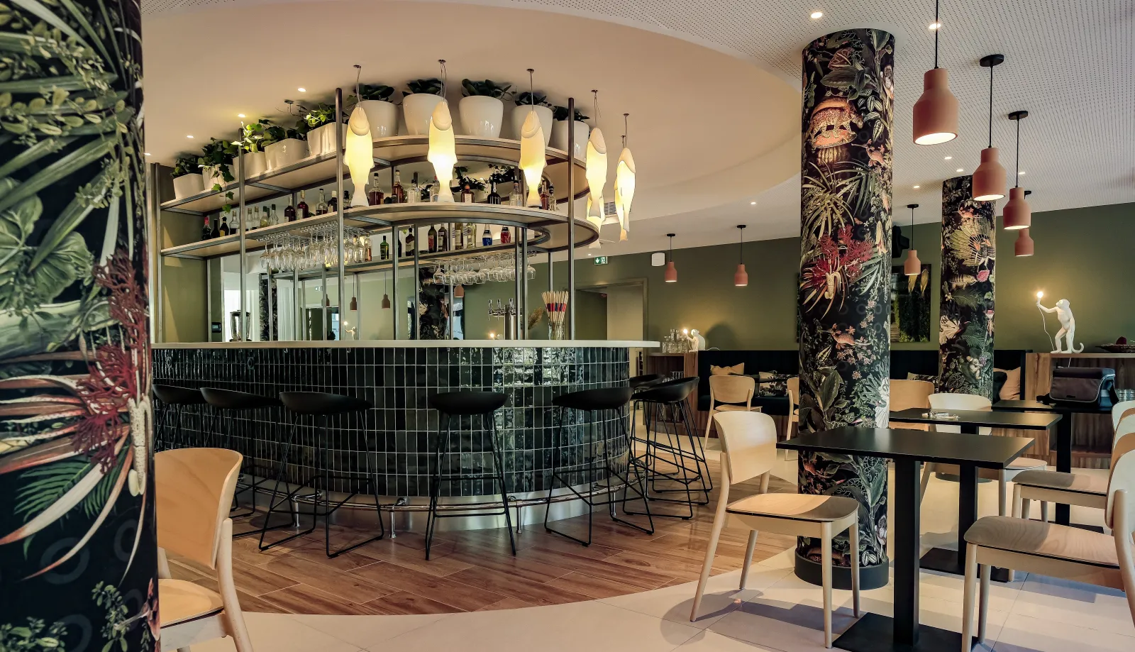 Modern hotel bar and lounge at Ibis Styles Bordeaux Lac Bruges featuring a curved black tiled bar counter with illuminated pendant lights, decorative botanical-patterned columns, comfortable seating areas, and potted plants throughout the space.