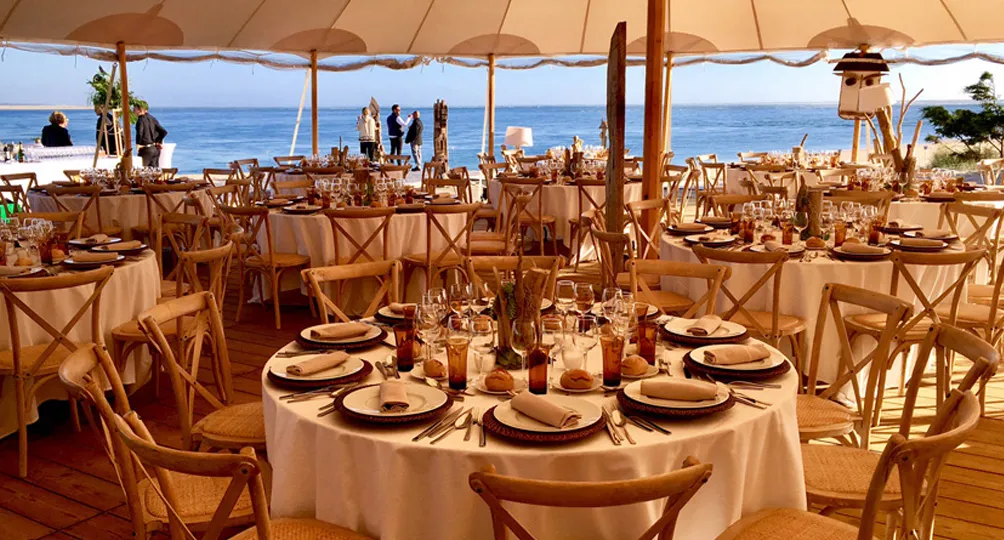 Elegant beachfront wedding reception setup with round tables dressed in white linens, wooden cross-back chairs, place settings, and sea view under tan umbrellas.
