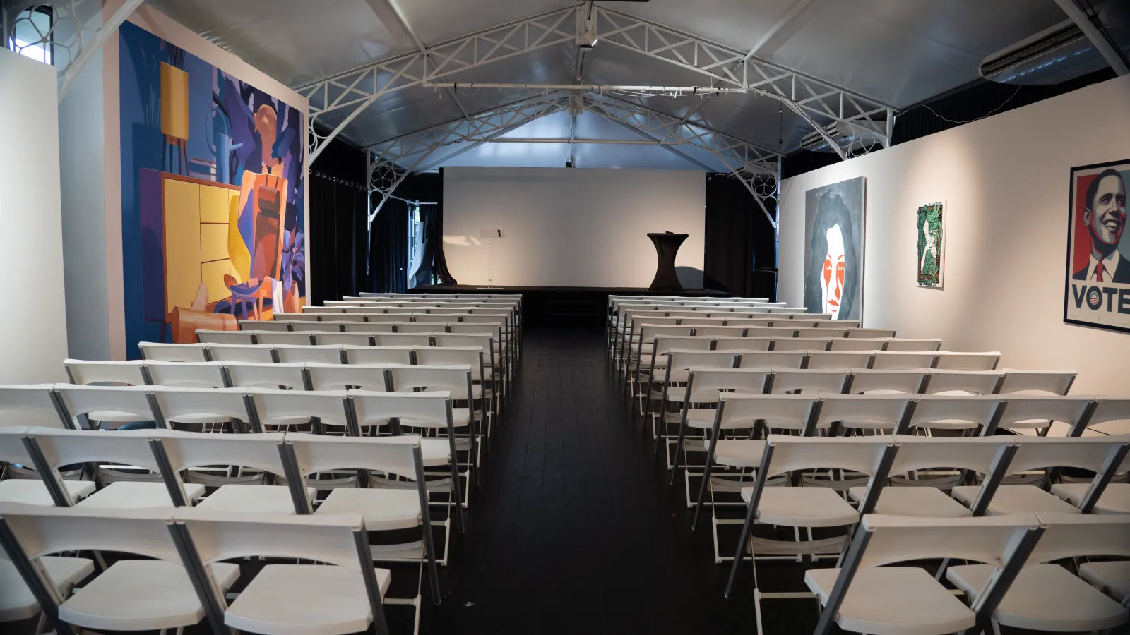 Interior view of an empty auditorium with white folding chairs arranged in rows facing a stage with a black podium, industrial ceiling with steel beams, and artwork displayed on the surrounding walls including colorful abstract paintings and political pos