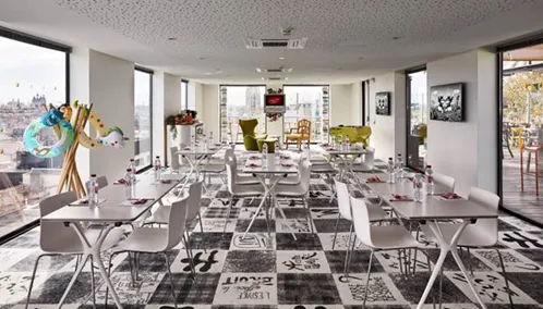 Modern dining room at Mama Shelter Bordeaux with long tables, contemporary chairs, geometric patterned flooring, floor-to-ceiling windows overlooking the city, and artistic decor elements.