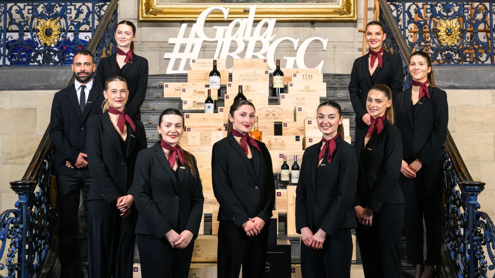 A professional team of nine staff members in formal black suits and burgundy ties posed on an elegant staircase in Bordeaux, with a large wine display featuring wooden boxes and bottles behind them, and ornate blue and gold decorative tiles visible on the