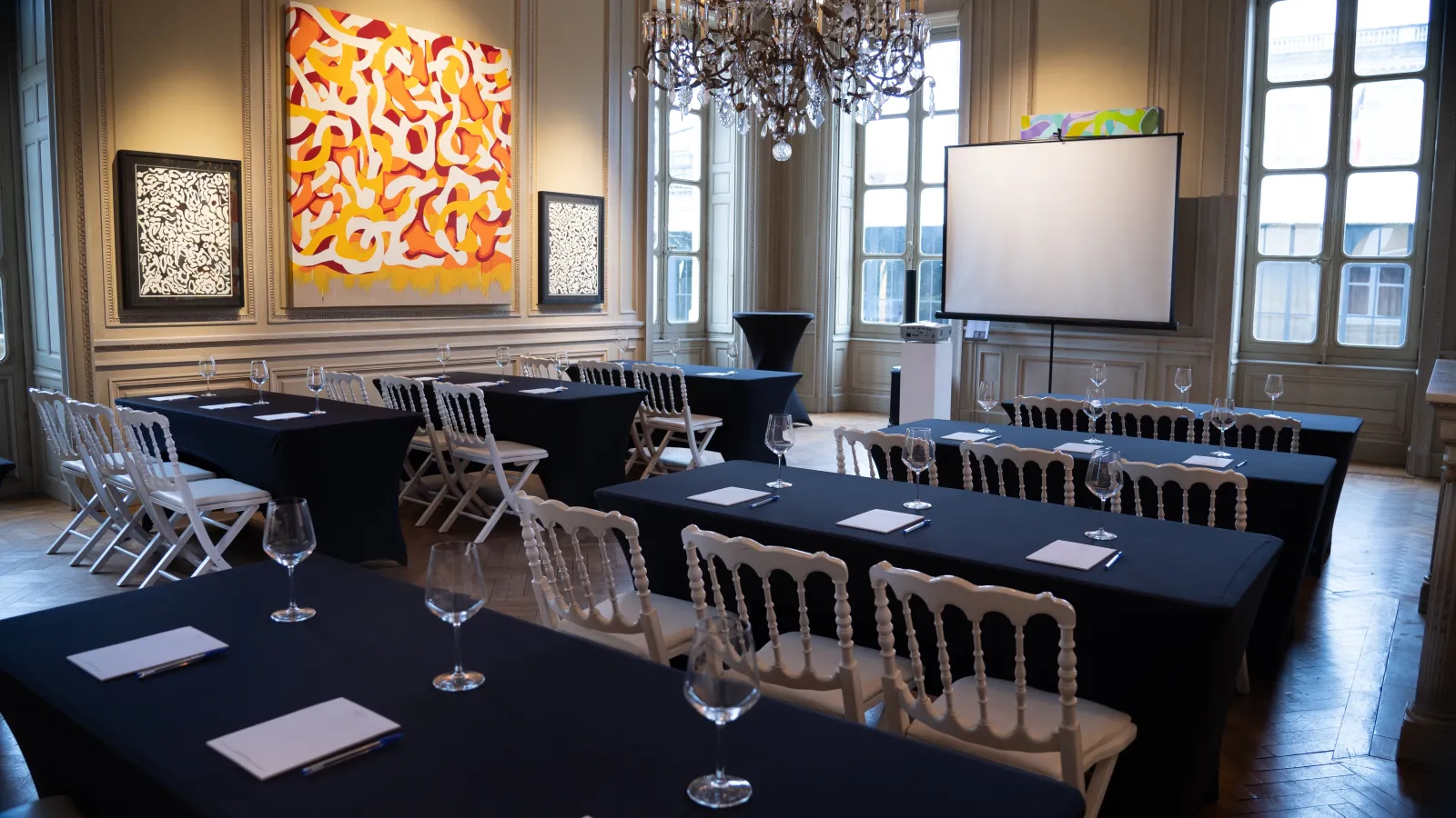 An elegant conference room at La Grande Maison de Bernard Magrez in Bordeaux, featuring long black tables with cream-colored chairs, a projection screen, crystal chandeliers, and contemporary artwork on beige walls with large windows.