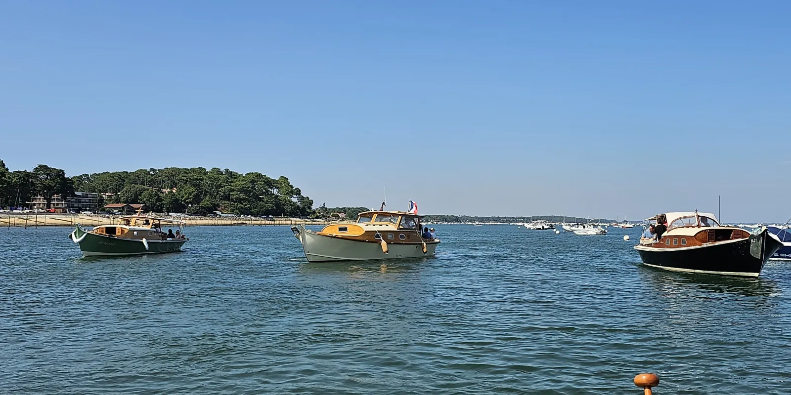 Moored fishing boats in a calm bay with a sandy beach and tree-lined shore in the background under a clear blue sky.