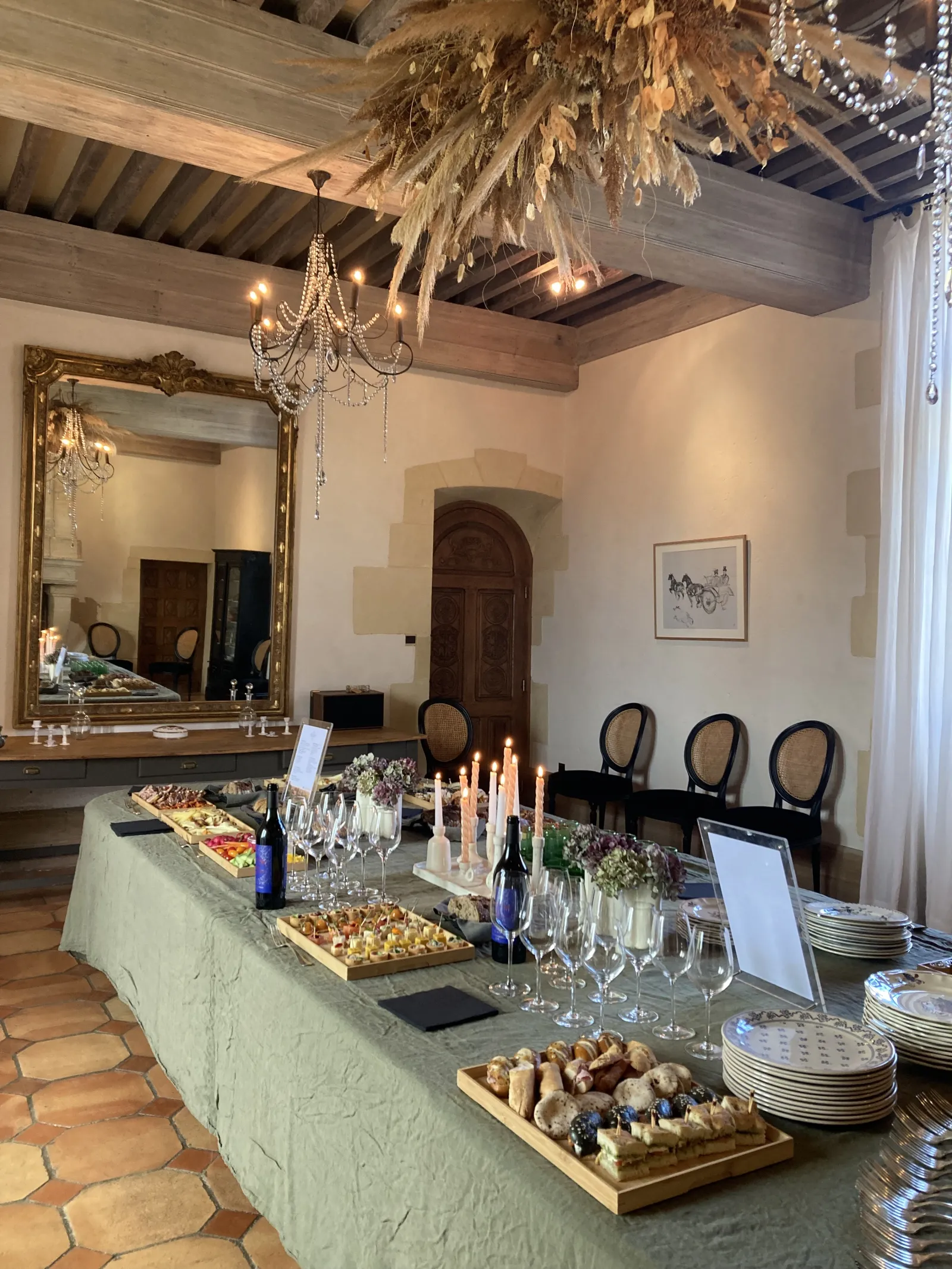 Elegant event space at Château Toulouse-Lautrec in Bordeaux featuring a marble buffet table with canapés, champagne flutes, and appetizers, ornate crystal chandelier, large gilt mirror, wooden beam ceiling, and formal seating