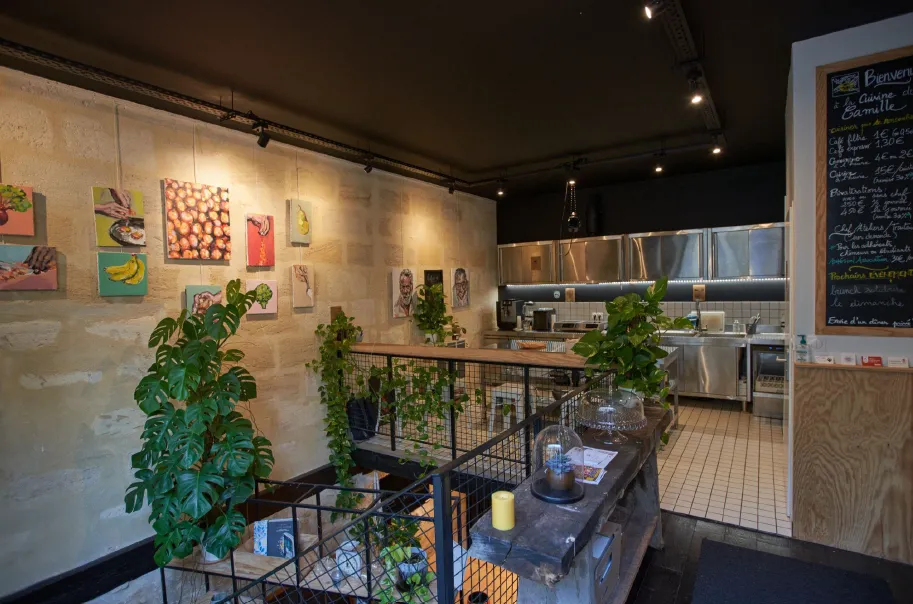 Interior of a modern restaurant kitchen and dining area with stainless steel equipment, displayed artwork on walls, green plants, and a chalkboard menu in Bordeaux