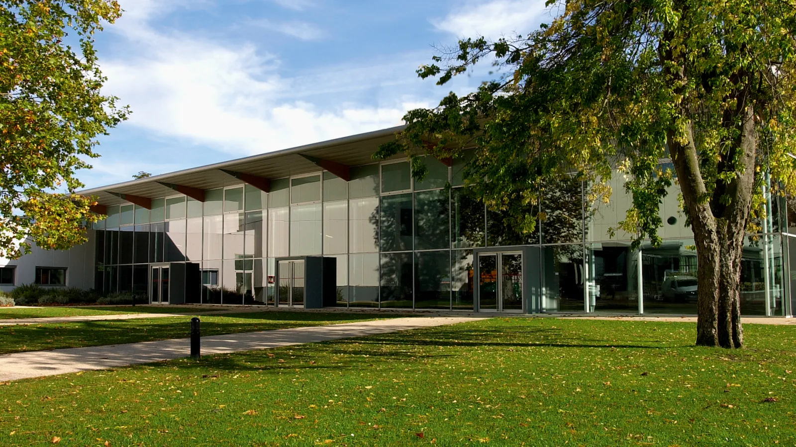 Modern architectural building with glass and white façade surrounded by green lawn and tall trees in autumn, located in Le Pin Galant, Bordeaux