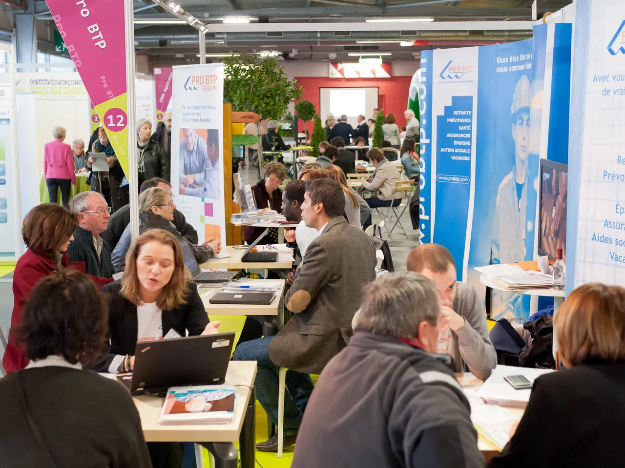 Busy trade fair or exhibition at Hangar 14 in Bordeaux, with numerous visitors consulting with exhibitors at booths displaying informational banners and materials about social services and insurance.