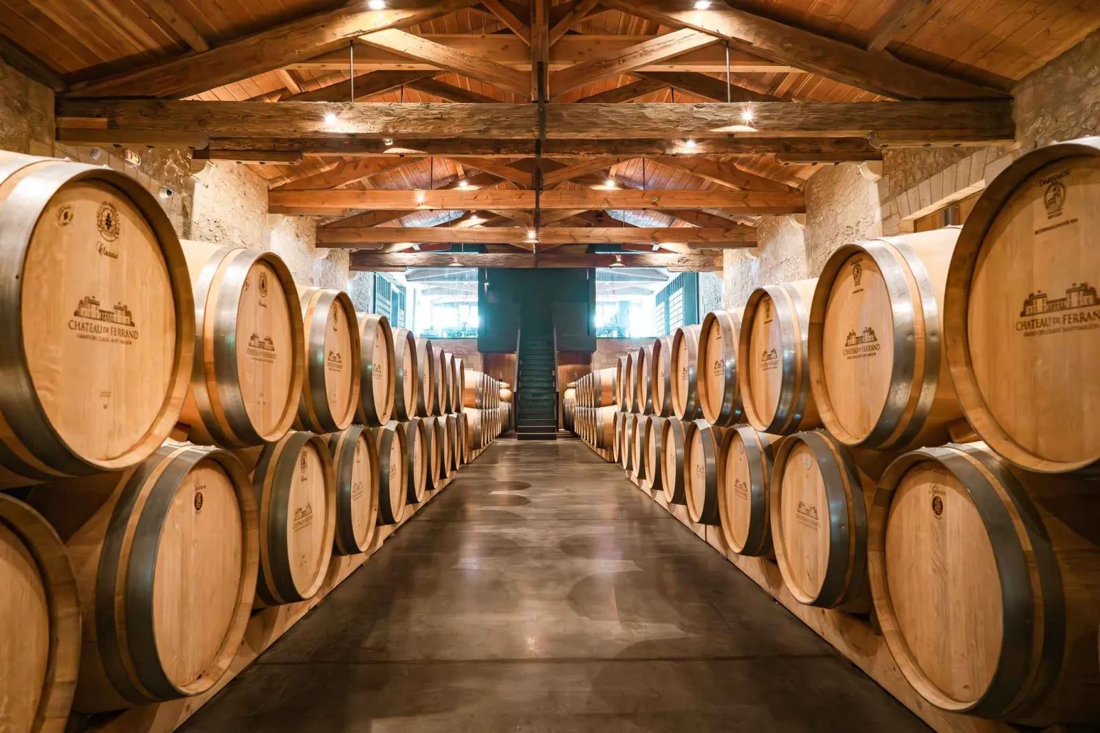 Wine barrel cellar at Château de Ferrand in Bordeaux, featuring rows of wooden oak barrels arranged symmetrically on both sides of a central corridor, with exposed wooden beam ceiling and stone walls.