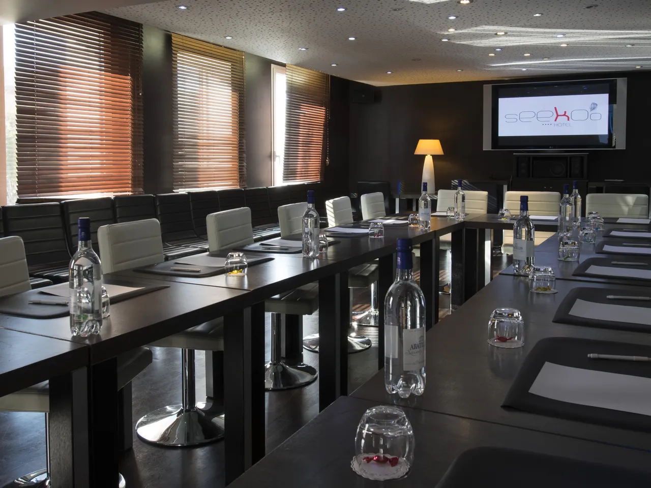 Modern conference room at Seeko'o Hotel Bordeaux with long black tables, cream-colored chairs, water bottles, notepads, and a presentation screen displaying the hotel logo. The room features orange-tinted blinds, recessed lighting, and floor-to-ceiling wi