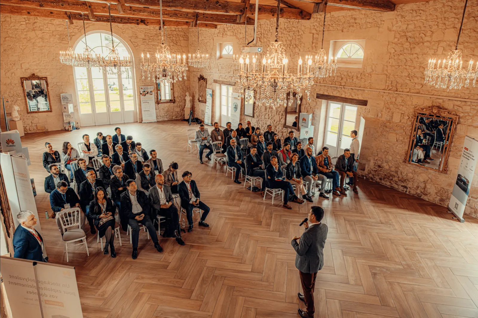 Aerial view of a professional conference or business event in an elegant reception hall at Château Gassies in Bordeaux, with attendees seated in a semi-circle facing a speaker, ornate chandeliers and arched windows visible