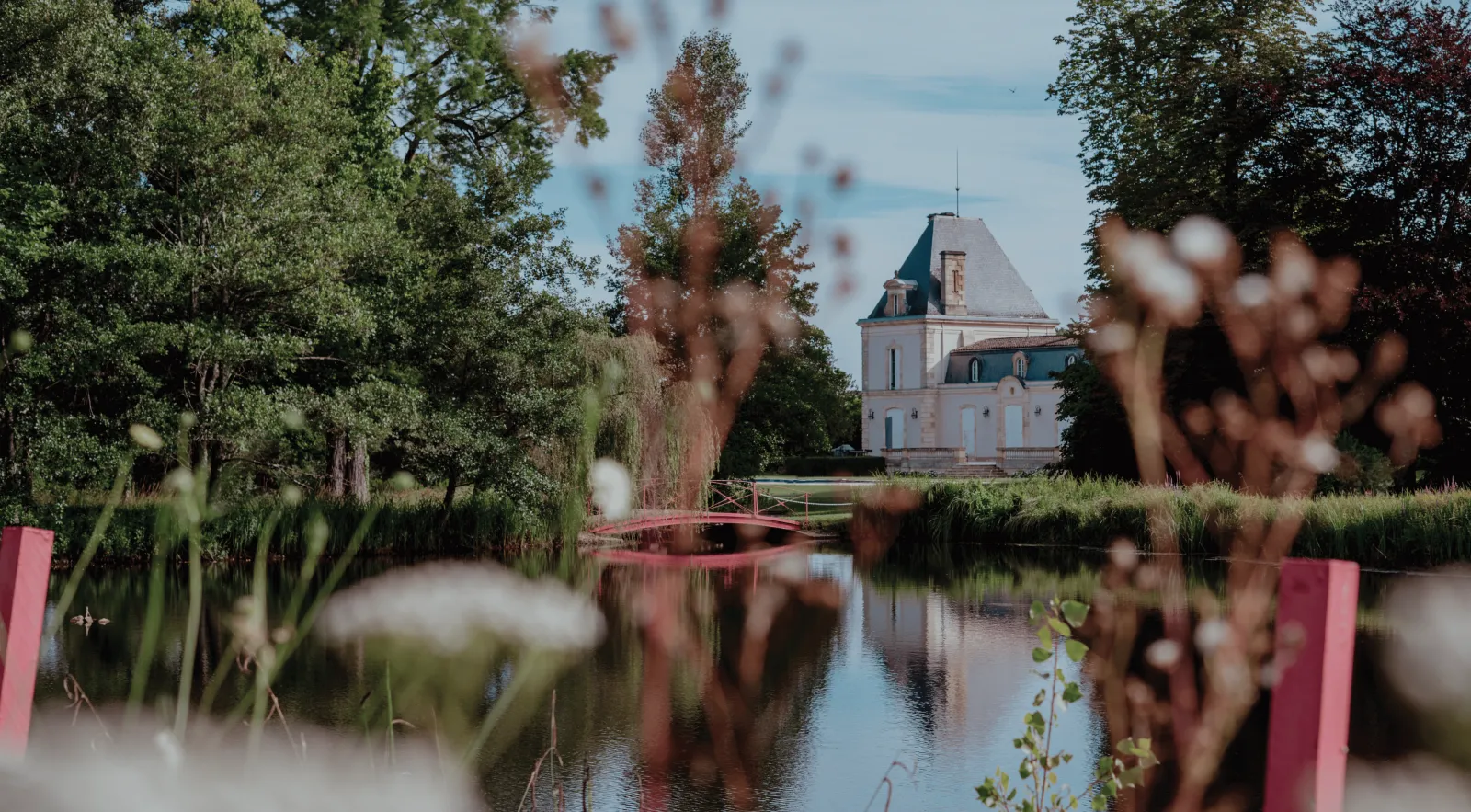 A picturesque view of Château Larrivet Haut-Brion in Bordeaux, featuring a pink arched bridge over a calm water canal, surrounded by lush gardens with blooming flowers, tall trees, and the château's distinctive mansard-roofed architecture visible in the b
