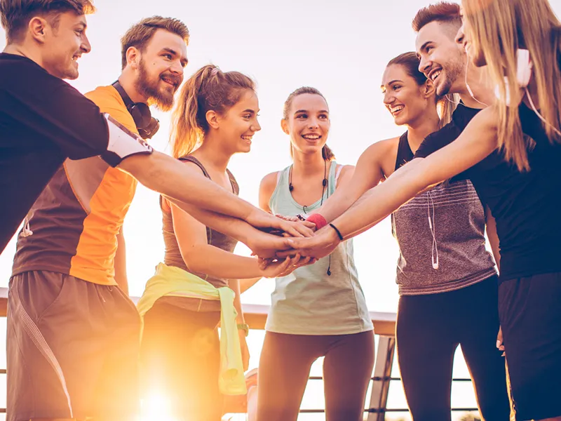 A diverse group of seven young people in sports attire putting their hands together in a circle, smiling and celebrating outdoors against a bright sky at sunset.