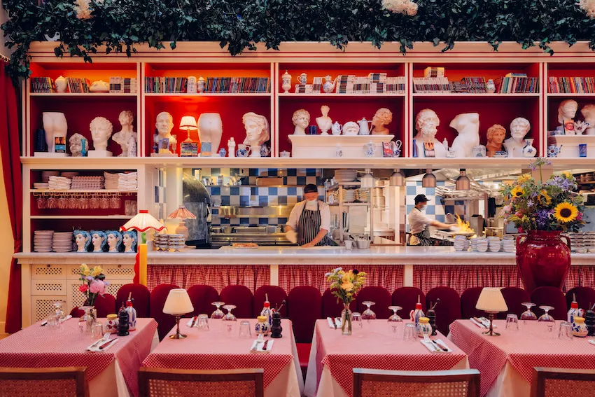 Interior of Osteria Palatino restaurant in Bordeaux featuring pink and red checkered tablecloths, burgundy velvet seating, open kitchen with blue tile backsplash, decorative busts and collectibles on shelves, and warm ambient lighting throughout the dinin