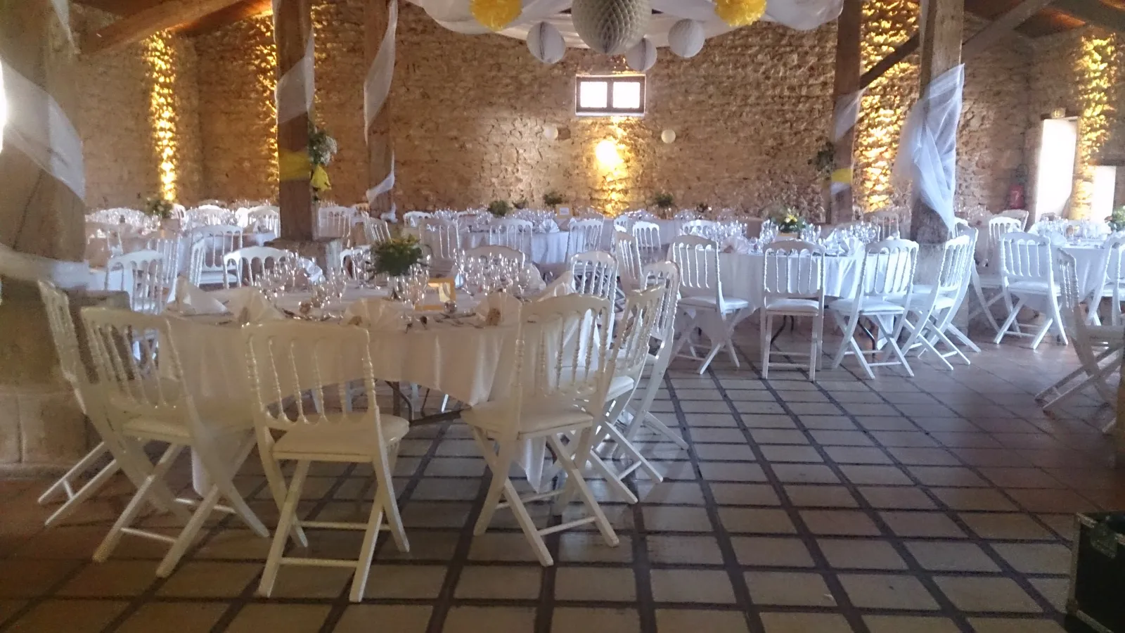 Elegant wedding reception venue in Bordeaux with white chairs arranged around tables with cream tablecloths, stone walls lit with warm golden lighting, and white decorative elements hanging from the ceiling.