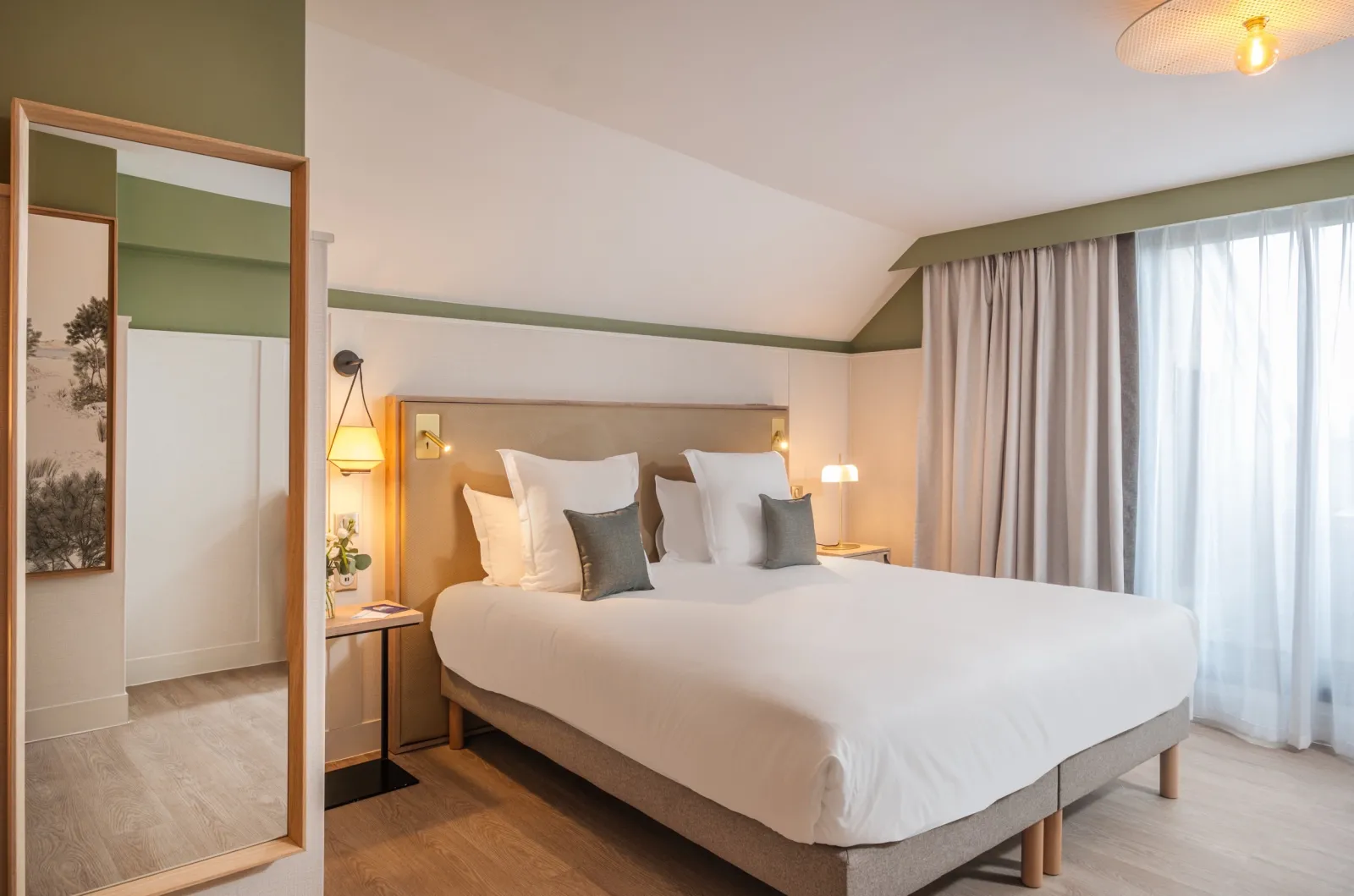 Modern hotel bedroom with a king-size bed, beige bedding, gray accent pillows, warm pendant lighting, green and white color scheme, wooden flooring, floor-to-ceiling curtains, and an open bathroom door visible on the left.