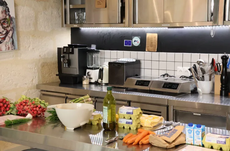 Modern kitchen countertop with fresh ingredients including red tomatoes, carrots, olive oil bottle, eggs, butter, and milk, with appliances and storage in the background at Camille's Kitchen in Bordeaux.