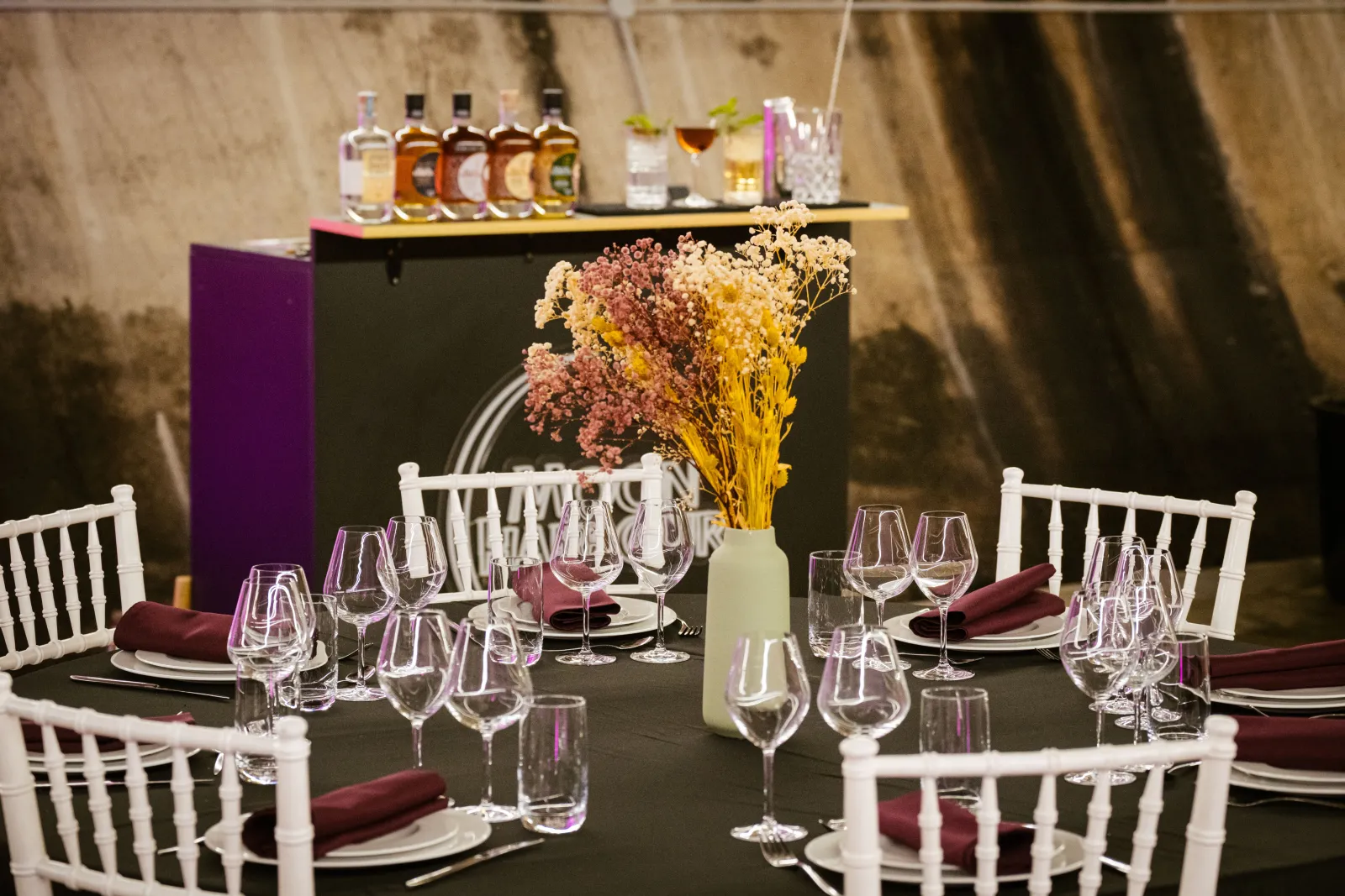 Elegant dining setup at Moon Harbour distillery in Bordeaux featuring a black table with wine glasses, burgundy napkins, white Chiavari chairs, dried flower centerpiece, and spirits bottles displayed on the bar in the background.